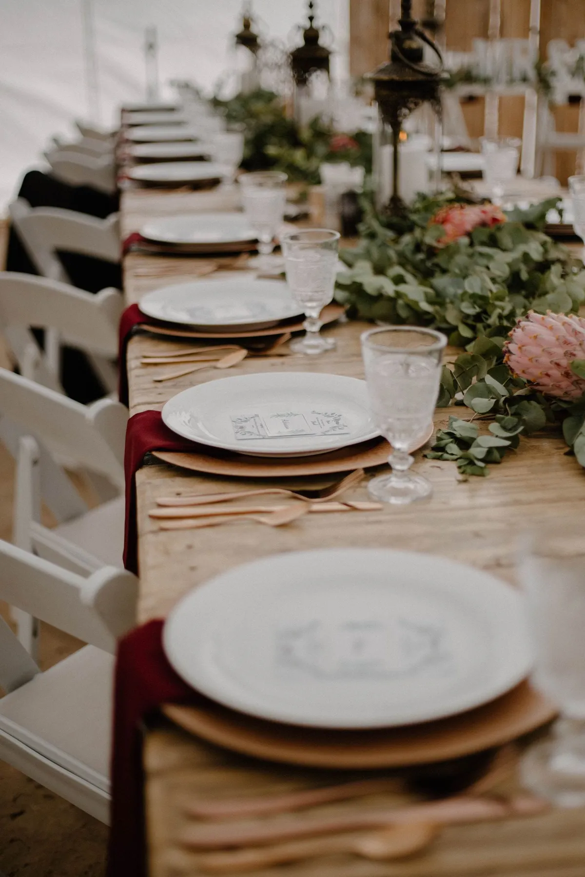 Mesa posta de casamento com guardanapos cor de vinho, folhagem de eucalipto e protea rosa sobre madeira